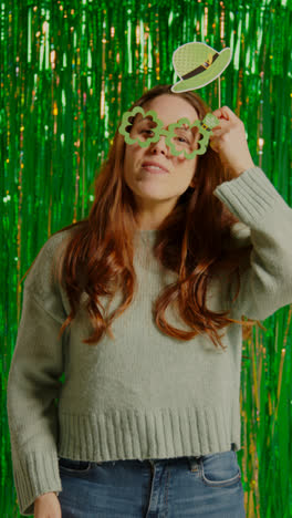 Vertical-Video-Of-Woman-Celebrating-St-Patrick's-Day-Standing-In-Front-Of-Green-Tinsel-Curtain-Wearing-Prop-Shamrock-Shaped-Glasses